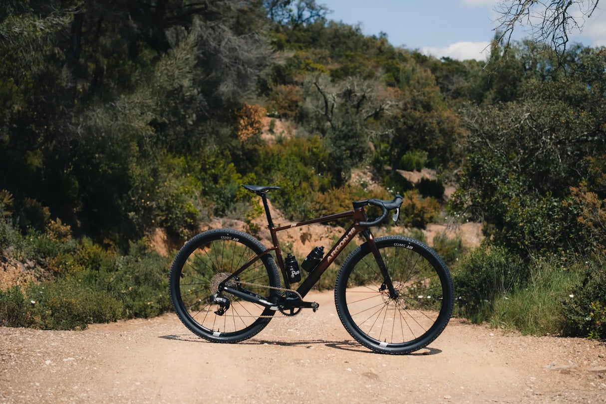 Argon 18 Dark Matter Rival XPLR AXS Gravel Bike parked on a dirt trail in a forest, showcasing its off-road capabilities and endurance design.