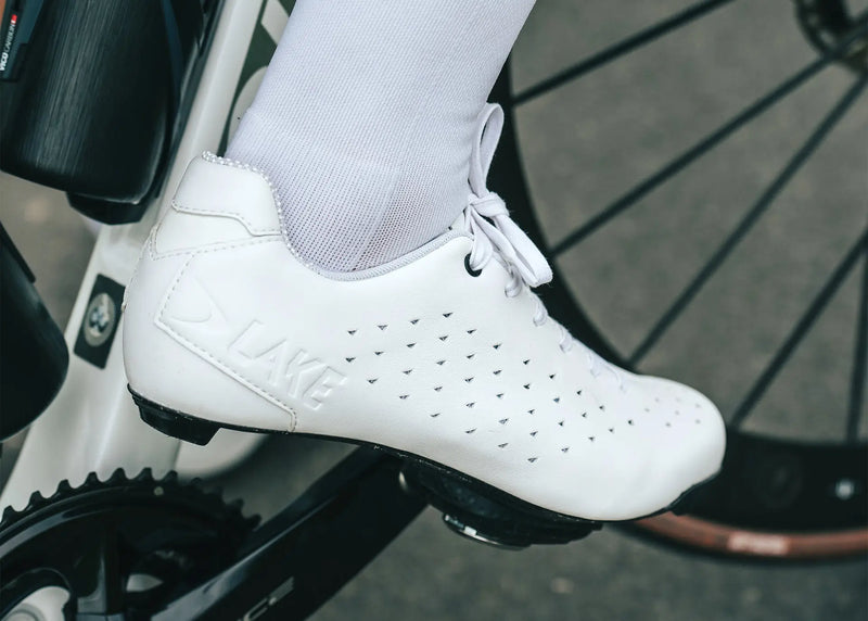 White cycling shoes with 'Lake' branding on a blurred bicycle background