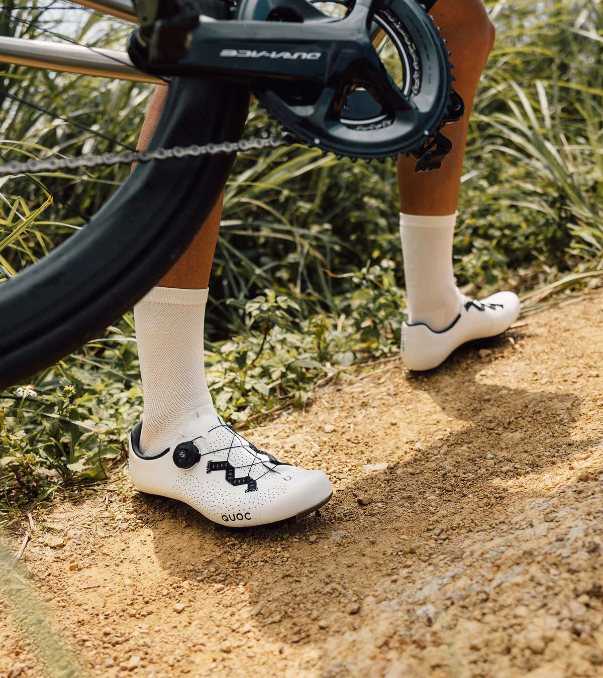 Cyclist walking on gravel trail wearing white QUOC Escape Road Shoes, ideal for adventure and endurance road rides.
