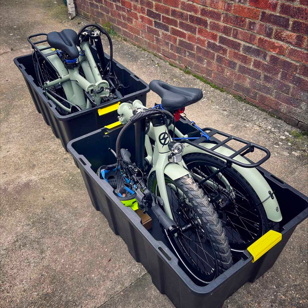 Two folded olive green estarli E20.8 Play Hybrid E-Bikes stored in black transport bins, ready for travel or storage.
