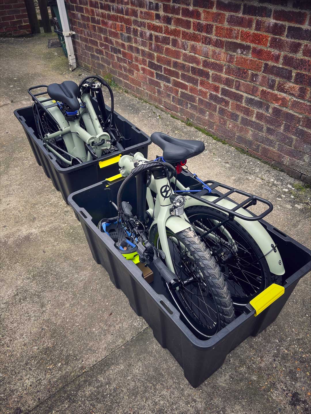 Two folded olive green estarli E20.8 Play Hybrid E-Bikes stored in black transport bins, ready for travel or storage.
