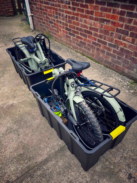 Two folded olive green estarli E20.8 Play Hybrid E-Bikes stored in black transport bins, ready for travel or storage.