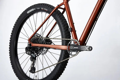 Close-up of the Cannondale Cujo 1 Kids Mountain Bike's rear section displaying the drivetrain, which includes the chain, gears, and rear wheel. The bike features a matte brown frame and thick, rugged black tires designed for off-road biking.