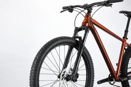 Close-up of the Cannondale Cujo 1 Kids Mountain Bike's front section on a white background. It showcases a matte orange frame, robust black suspension fork, disc brake, and thick treaded tire. The handlebars and part of the seat are visible.