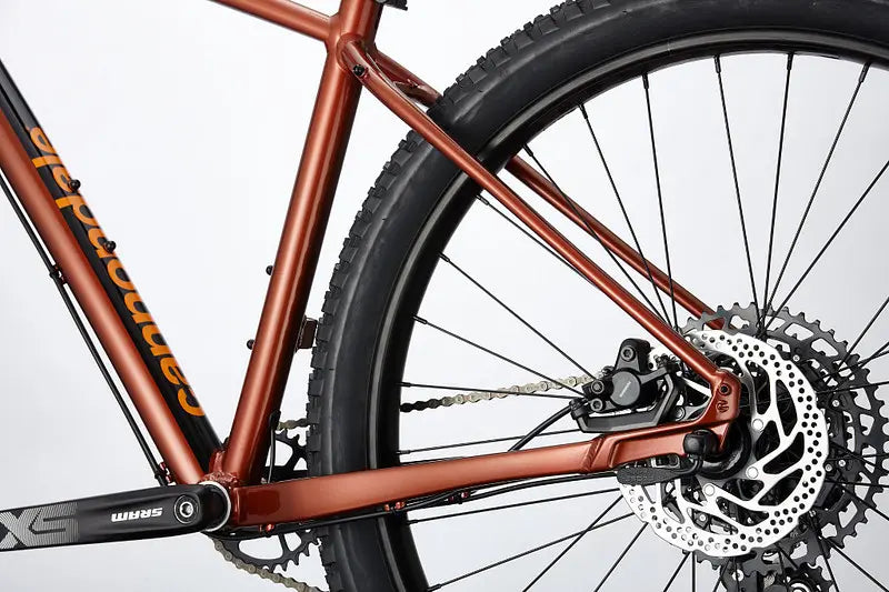 Close-up of the rear wheel and frame of the Cannondale Cujo 1 Kids Mountain Bike. The bike showcases a metallic orange frame with disc brakes featuring a detailed silver rotor and a black tire. The Cannondale brand logo is visible on the frame against a plain white background.