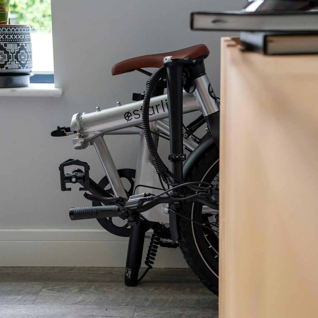 Folded silver estarli E20.8 Play Hybrid E-Bike stored indoors beside a desk, showcasing its compact design for home storage.