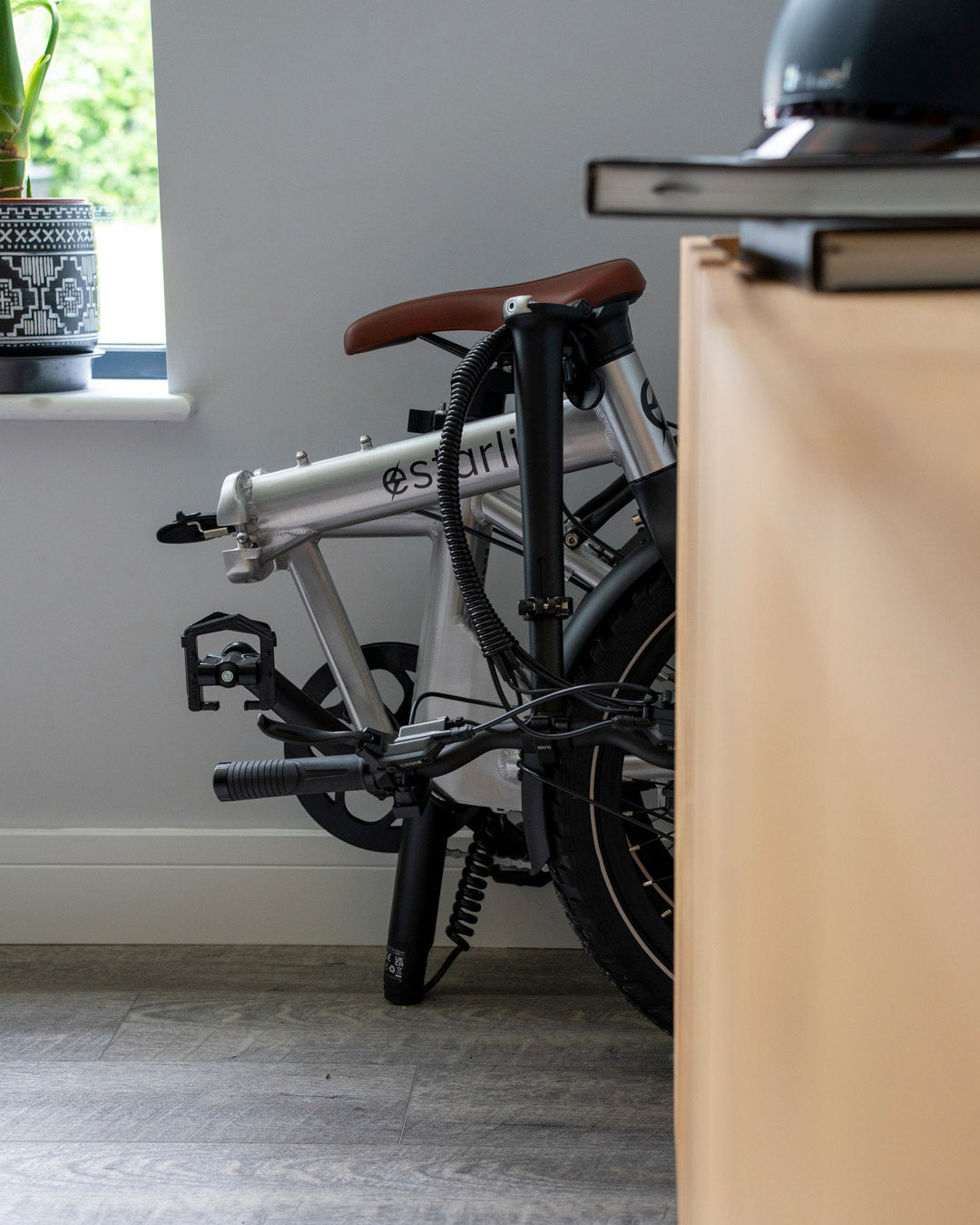 Folded silver estarli E20.8 Play Hybrid E-Bike stored indoors beside a desk, showcasing its compact design for home storage.