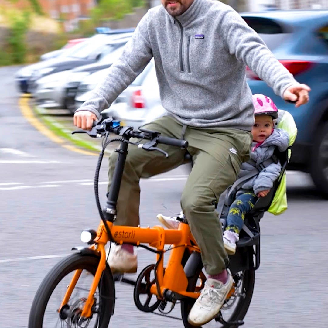 Man riding orange estarli E20.7 Comfort Hybrid E-Bike with child seat, showcasing family-friendly design and urban mobility.