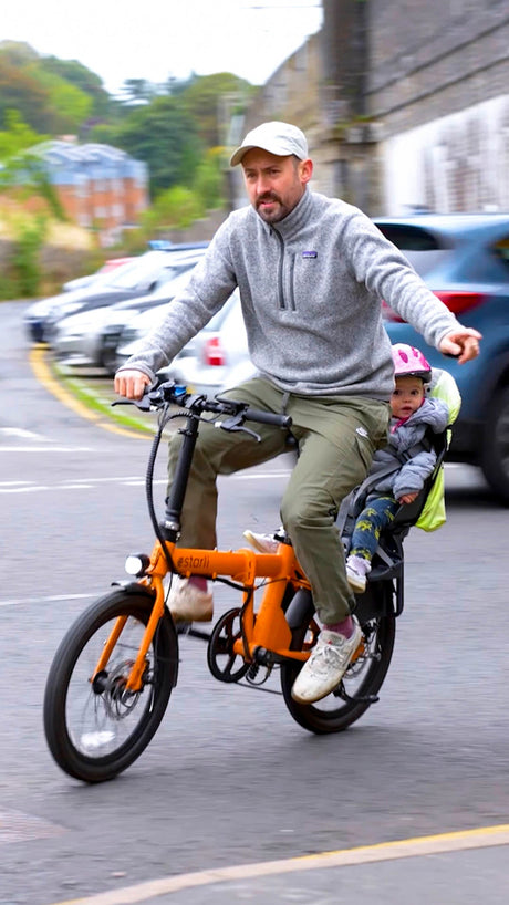 Man riding orange estarli E20.7 Comfort Hybrid E-Bike with child seat, showcasing family-friendly design and urban mobility.