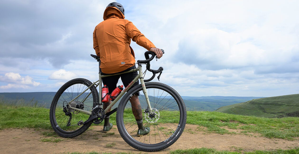 Cyclist standing with the 2025 Genesis Croix De Fer 30 Gravel Bike on a scenic hilltop, ready for an off-road adventure ride.