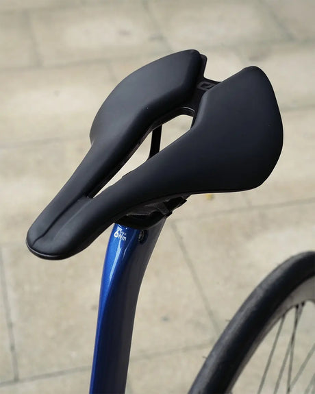 A modern, sleek black bicycle saddle on a blue seat post blends seamlessly with the Pinarello Dogma F Disc Ult 12 Di2 Campagnolo Shamal Road Bike against a blurred stone-tiled background. The saddle boasts a unique, aerodynamic design for enhanced comfort and performance.