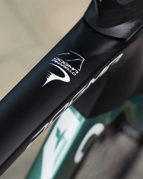 Close-up view of a Pinarello Dogma F Ultegra 12 Di2 Racing Zero road bike frame in black, showcasing a prominent white logo design with a stylized helmet and the uppercase "PINARELLO" text. The frame boasts aerodynamic lines and a smooth finish, enhanced by the precision engineering of its Ultegra Di2 components.