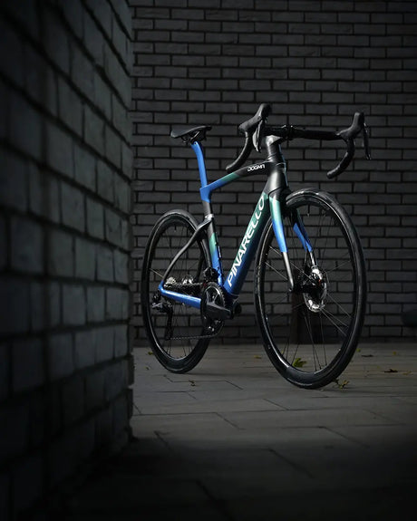 A sleek, modern Pinarello Dogma F Disc Ult 12 Di2 Shamal Road Bike in blue and black stands against a backdrop of gray brick walls. The bike is well-lit, highlighting its streamlined design and high-performance features, complemented by Shamal wheels for an exceptional ride.