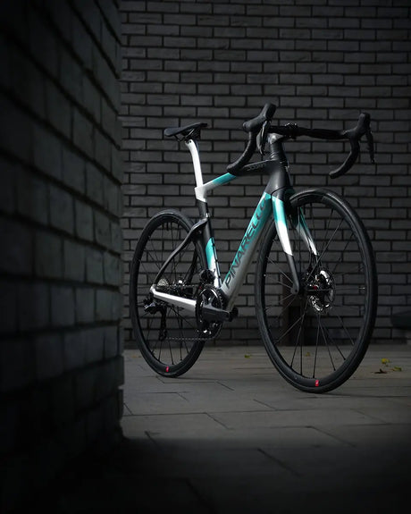 A sleek, modern Pinarello Dogma F Ultegra 12 Di2 Racing Zero road bike with a turquoise and white frame is positioned against a dark brick wall. The lighting casts a shadow on the pavement, highlighting its streamlined design, Ultegra Di2 drivetrain, and black components.