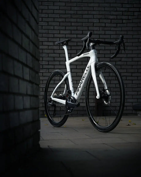 A sleek, white Dogma F Disc Ult 12 Di2 Campagnolo Shamal Road Bike with black handlebars and wheels leans against a dark brick wall. The dim lighting perfectly highlights this modern road bike's design and the Pinarello brand name on its frame.