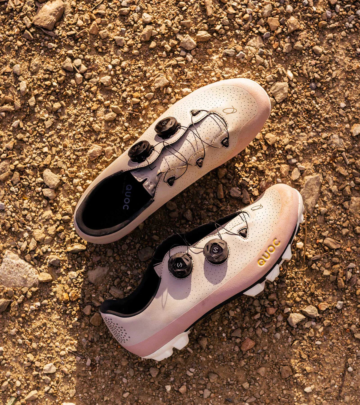 Pair of QUOC Gran Tourer XC Off-Road Shoes in sand and pink resting on gravel, showcasing durable design for all-terrain cycling.