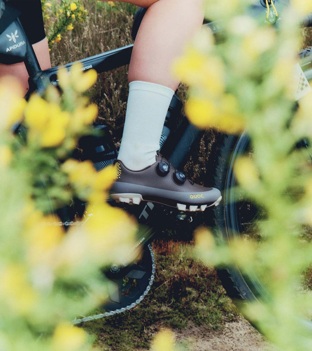 Cyclist wearing QUOC Gran Tourer XC Off-Road Shoes in brown, riding through a trail surrounded by yellow flowers, emphasizing outdoor use.