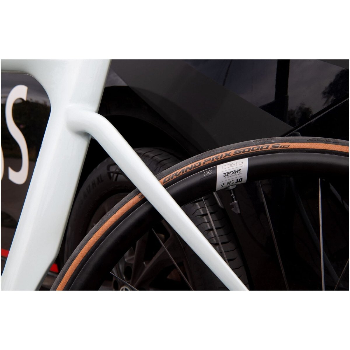 Close-up shot of the rear wheel and frame of a white bicycle, positioned beside a black car. The tire reads "Grand Prix 5000" and showcases detailed treads. The image highlights the intersection of the bike's structure and the car's surface, featuring a DT Swiss A 1800 SPLINE disc brake wheel, clincher 30 x 20 mm, at the front.