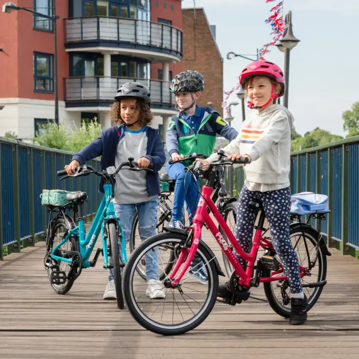 Three children, each wearing a helmet, stand with their Frog City 61 Kids Hybrid Bikes on a wooden bridge. Their bikes, featuring lightweight frames and 8-speed gears, suggest they're prepared for an adventure. With buildings and greenery in the background, the bright day promises pleasant weather for their ride.