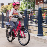 A young child wearing a pink helmet and backpack rides a Frog City 61 Kids Hybrid Bike with 8-Speed Gears on a paved path. The child is dressed in a striped sweater and polka dot pants. A railing and greenery are visible in the background, perfectly complementing the bike's lightweight frame.