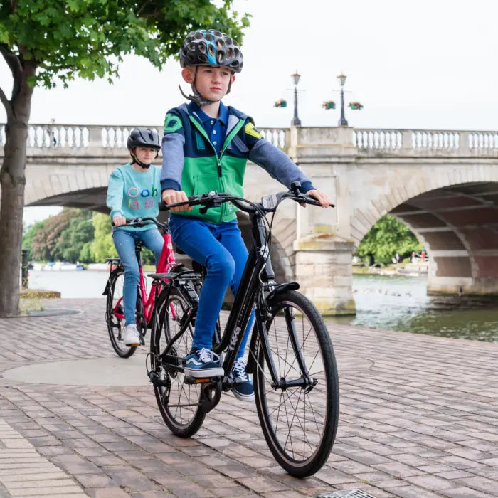 Two children on Frog City 61 Kids Hybrid Bikes ride along a riverside path, both wearing helmets and casual attire. Their lightweight frame bikes with 8-speed gears ensure a smooth journey as they pass by a stone bridge and trees, with the river gently flowing below.