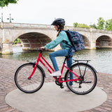 A person wearing a helmet and carrying a large backpack stands next to their Frog City 61 Kids Hybrid Bike by the riverside. The red bicycle, from the brand Frog, is equipped with 8-speed gears. In the background, a stone bridge can be seen, while trees flank the paved pathway along the river.