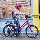 A child wearing a pink helmet and backpack confidently rides their vibrant pink Frog City 61, a kids hybrid bike with a lightweight frame. Dressed in polka dot leggings and a light jacket, they pedal past a decorative shop window adorned with string lights and a yellow pillar.