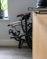 Folded silver estarli E20.8 Play Hybrid E-Bike stored indoors beside a desk, showcasing its compact design for home storage.