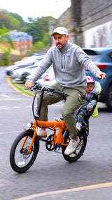 Man riding orange estarli E20.7 Comfort Hybrid E-Bike with child seat, showcasing family-friendly design and urban mobility.