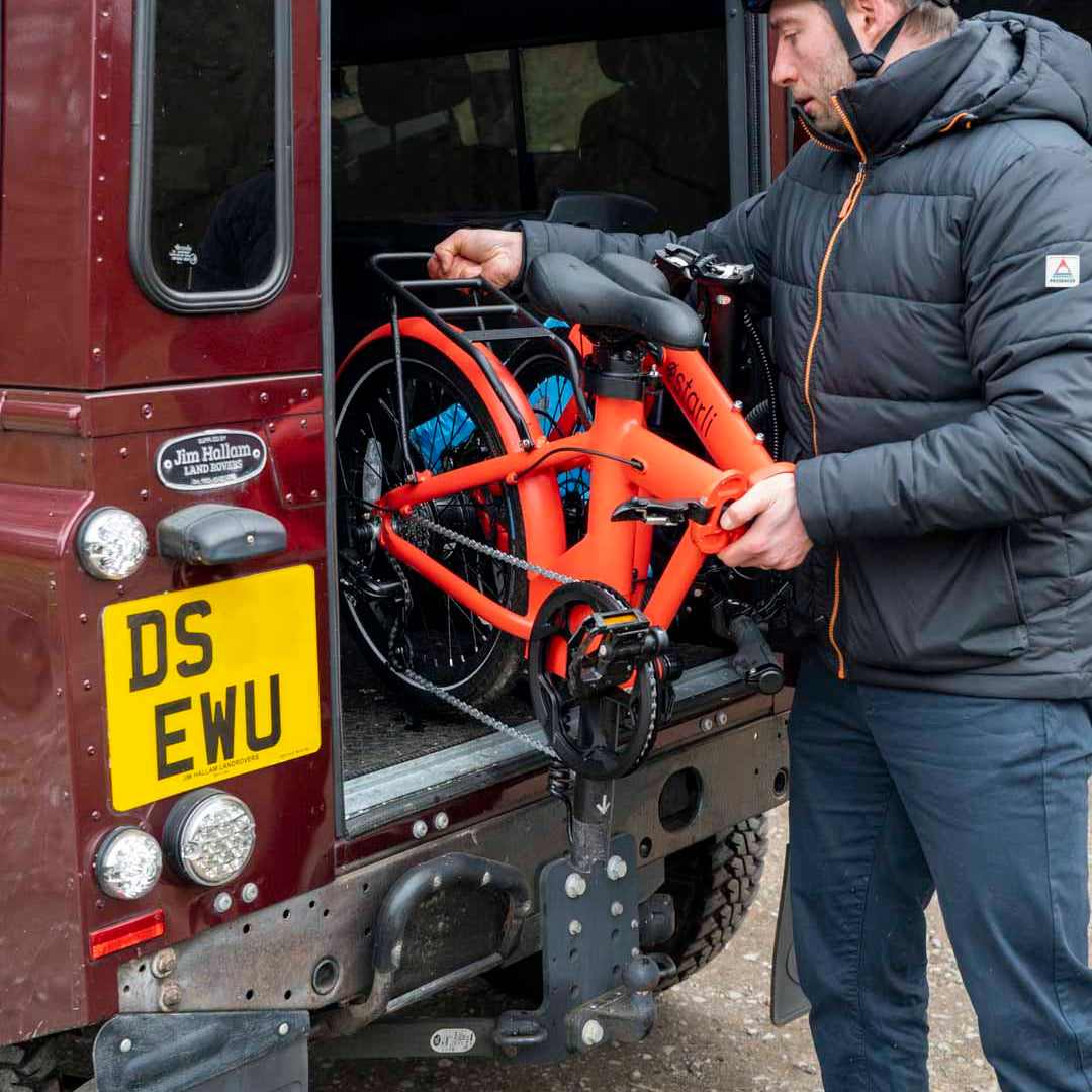 Man loading folded orange estarli E20.7 Comfort Hybrid E-Bike into SUV, highlighting its compact portability for travel and storage.