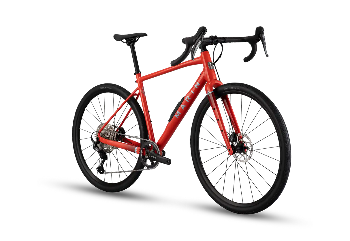 A red gravel bicycle with black components, drop handlebars, and a black saddle, shown from a three-quarters angle on a white background with a soft shadow.