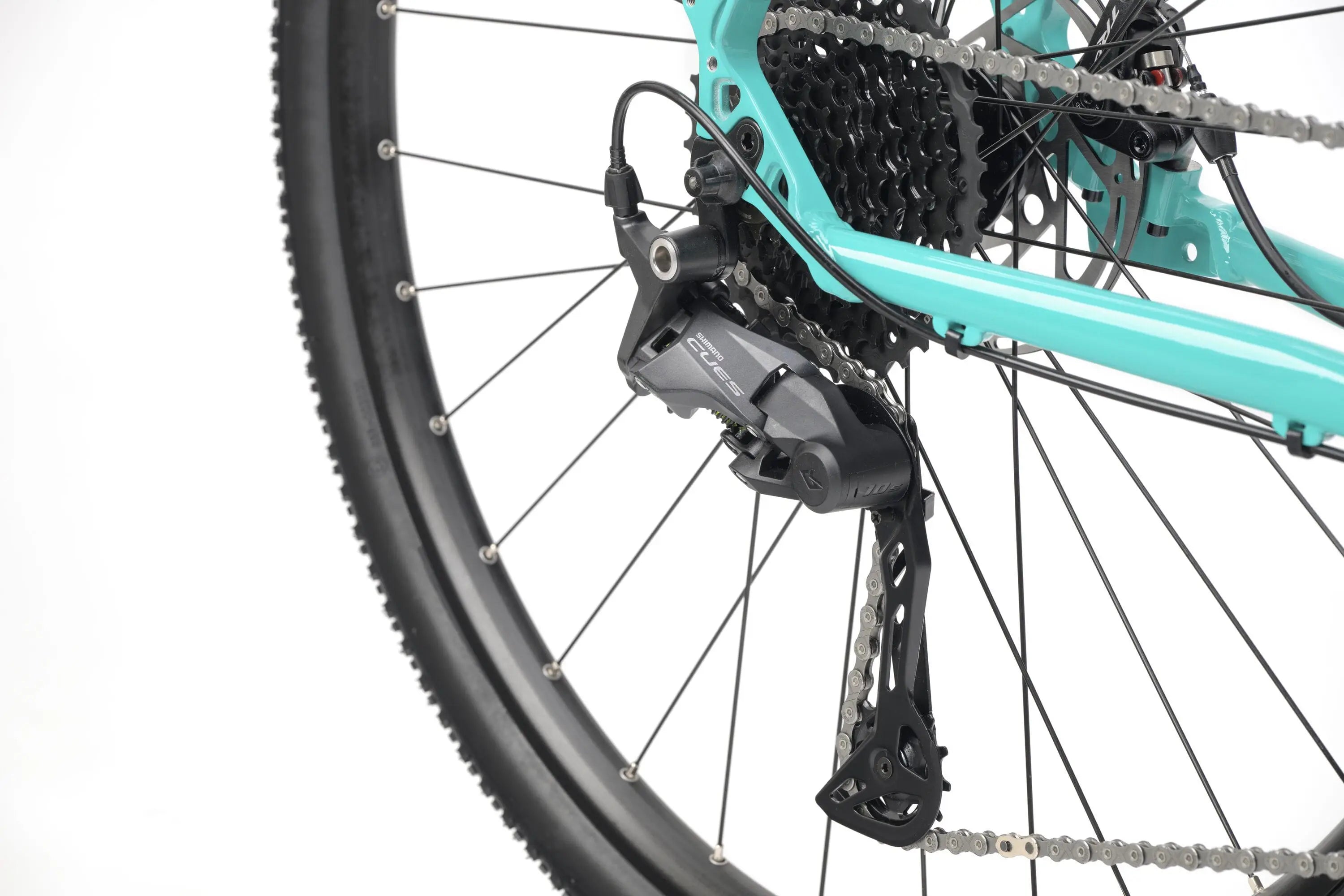 Close-up of a teal gravel bike rear derailleur and cassette with knobby tire and black spokes.