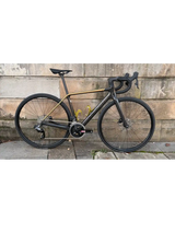 Matte black Cervelo R5 Rival AXS road bike with gold accents, carbon frame, and drop handlebars against a stone wall.