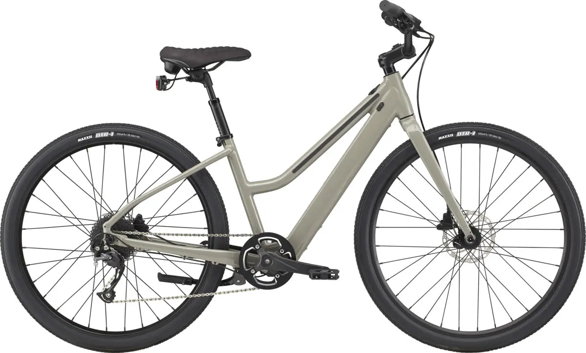 The Cannondale Treadwell Neo Remixte Electric Hybrid Bike, crafted by Cannondale, is a stylish gray E-bike boasting a minimalist design. It features a step-through frame, thick black tires, and a rear light. Its lightweight battery delivers a smooth ride, while the suspension seat post and flat handlebars provide comfort for urban commuting.